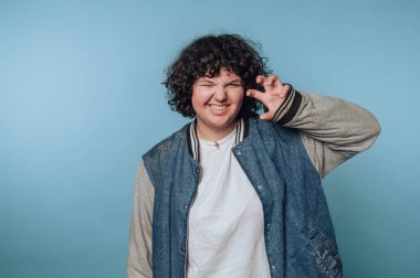 Kıvırcık saçlı, kot ceket giyen ve beyaz gömlekli biri mavi arka plana karşı neşeli bir jest yapar..