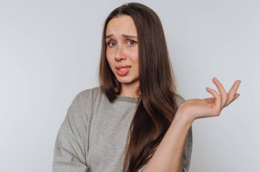 Uzun kahverengi saçlı ve gri kazaklı bir kadın karmaşık bir ifade takınır. Bir elini kaldırır ve basit bir arka plana doğru sorgulama hareketi yapar..