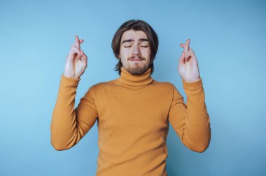 Uzun saçlı ve sakallı, balıkçı yaka kazak giyen genç bir adam mavi bir arka plana karşı duruyor gözleri kapalı ve parmakları çapraz, umut ya da hüsnükuruntu iletiyor.