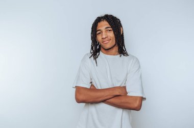 Beyaz tişörtlü örgülü genç adam, kollarını kavuşturmuş, gülümsüyor..