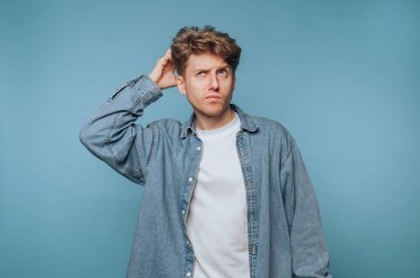 Saçları dağınık, beyaz bir tişörtün üzerine kot ceket giyen genç bir adam açık mavi bir arka planda duruyor. Düşünceli görünüyor. Kafasını karıştırıyor. Şaşırmış..