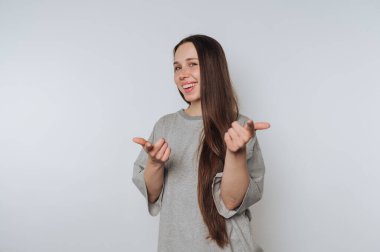 Uzun kahverengi saçlı, gri tişörtlü, neşeli bir şekilde kameraya gülümseyen ve beyaz bir arka plana bakan genç bir kadın..