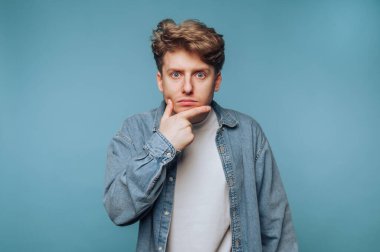 Düşünceli bir yüzü olan genç bir adam, kot gömlek giyiyor, mavi bir arka planı var..