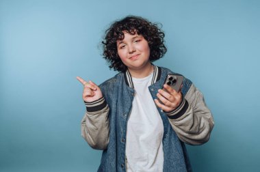 Kıvırcık saçlı, kot ceket giyen bir adam elinde bir akıllı telefon tutuyor, mavi arka planı işaret ediyor..