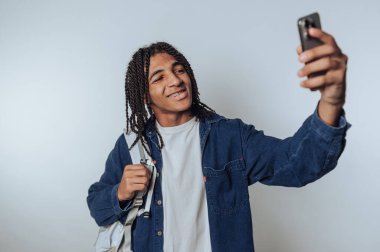 Örgülü saçlı genç adam selfie çekiyor, kot ceket giyiyor ve sırt çantası tutuyor, arka planda sırıtıyor..