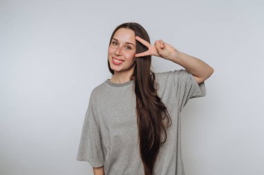 Uzun kahverengi saçlı, gri tişörtlü, gülümseyen ve beyaz arka planda barış işareti yapan genç bir kadın..