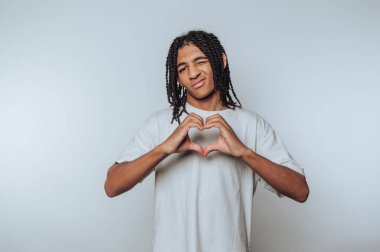 Saçları örgülü genç bir adam elleriyle kalp şekli yapıyor açık gri bir arka plana neşeyle gülümsüyor..
