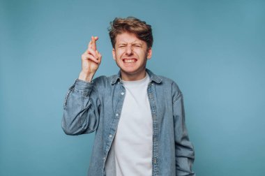 Kıvırcık saçlı genç bir adam mavi bir arka plana karşı umut ya da beklenti içinde parmaklarını ve gözlerini kapatır..