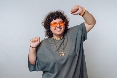 Kıvırcık saçlı, turuncu güneş gözlüğü takan, bol gri tişörtlü, açık bir arkaplanda neşeyle dans eden neşeli bir insan..