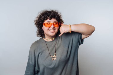 Kıvırcık saçlı, turuncu güneş gözlüğü takan ve gri tişörtlü biri elleriyle telefon hareketi yapıyor. Renkli bilezikler takıyorlar ve kolyelerinde dinozor kolyesi var..