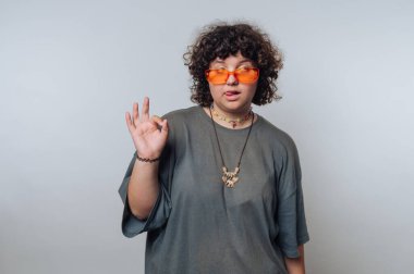 Kıvırcık saçlı, turuncu güneş gözlüklü ve gri gömlekli biri el hareketi yapıyor. Arka plan açık gri, modern ve sıradan bir hava yaratıyor..