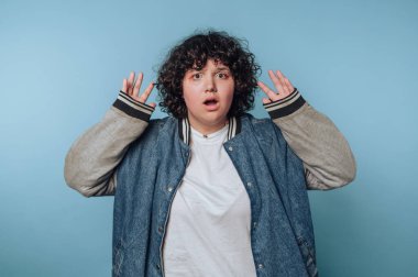 Kıvırcık saçlı, mavi arka planda kot ceket giyen şaşırmış biri..
