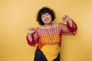 Kıvırcık saçlı, sarı gömleğin üstüne kırmızı ağ takan neşeli bir kadın sarı arka plana karşı duruyor, gülümsüyor ve kendini işaret ediyor..