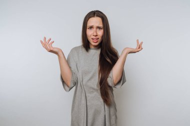 Gri tişörtlü, uzun saçlı genç bir kadın düz bir arkaplan karşısında duruyor. Karışıklığı ya da inanmazlığı ifade ediyor. Kolları havada ve avuç içleri havada..
