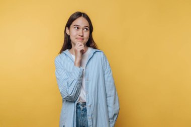 Mavi çizgili gömlekli düşünceli genç bir kadın sarı arka planda poz veriyor..