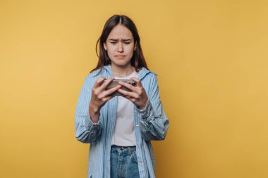 Genç bir kadın, akıllı telefonunu sarı arka planda kullanırken hayal kırıklığına uğramış görünüyor..