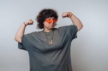 Kıvırcık saçlı ve turuncu güneş gözlüklü biri kaslarını düz bir arkaplana yaslar..