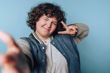 Kıvırcık saçlı, kot ceket giyen ve beyaz gömlekli biri, neşeli bir poz veriyor, göz kırpıyor ve mavi arka plana barış işareti yapıyor..