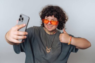 Turuncu güneş gözlüğü takan kıvırcık saçlı bir insan akıllı telefonuyla selfie çeker ve olumlu bir jest yapar. Arka plan sade ve insan gülümsüyor..