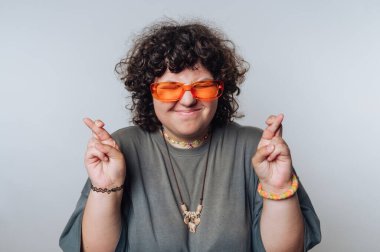 Kıvırcık saçlı, turuncu güneş gözlüklü ve gri gömlekli umut dolu bir ifadeyle parmaklarını çapraz yapan biri..