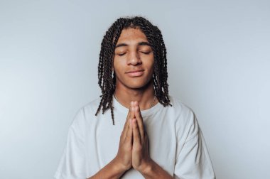 Saçları örgülü, gözleri kapalı beyaz bir gömlek giyen genç bir adam, sade bir arka plana karşı bir dua jestinde el ele tutuşuyor..