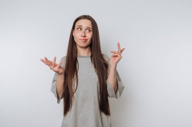 Uzun kahverengi saçlı, gri tişörtlü, kafası karışık bir ifadeyle düz beyaz bir arka plana karşı omuz silken genç bir kadın..