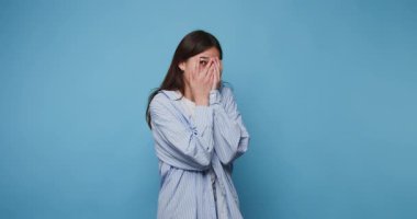 Genç bir kadın, bir stüdyoda sakin bir anda yüzünü mavi bir arka planda gizlerken duygularını ifade eder.