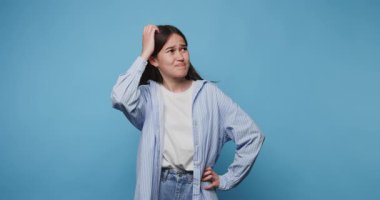 Gündelik kıyafetli genç kadın mavi arka planda kafa karışıklığı yaratıyor.