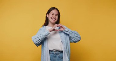 Genç bir kadın, elleri parlak sarı bir arka planda neşeyle kalp şekli oluşturuyor.