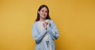 Gülümseyen genç bir kadın, parlak sarı bir arka plana karşı kalp hareketi yapıyor.