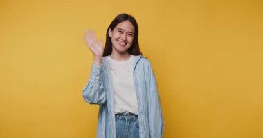 Gülümseyen genç bir kadın açık sarı bir arkaplan önünde el sallıyor.