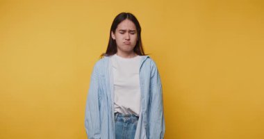 Genç kadın, günlük kıyafetlerini giyerken parlak sarı arka plana karşı kafa karışıklığı gösteriyor.