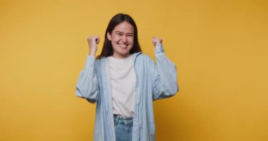 Neşeli genç kadın, parlak sarı arka planda mutlu bir şekilde dans ediyor ve zaferle duygusal olarak seviniyor.