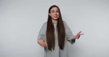 Gri tişörtlü kafası karışmış bir kadın beyaz arka planda kollarını yana açıyor ve kafasını karıştırarak kameraya bakıyor.