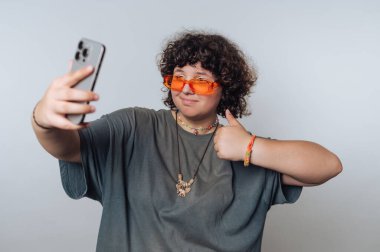 Şaşırmış kıvırcık saçlı genç bir kadının portresi selfie çekmekten, şık turuncu gözlükleriyle gösteriş yapmaktan ve açık arkaplanda güzel bir jest yapmaktan hoşlanıyor.