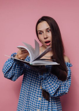 Mavi kareli gömlekli genç bir kadının portresi pembe arkaplan üzerine bir kitap okurken tepki veriyor