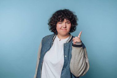 Genç bir kadının portresi, şakacı bir ifadeyle parmağını kaldırıyor, mavi arka planda eğlendirici ve spontane bir şekilde.