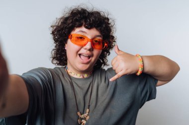 Şaşırmış kıvırcık saçlı genç bir kadının portresi açık arkaplanda neşeli bir jestle selfie için poz veriyor.