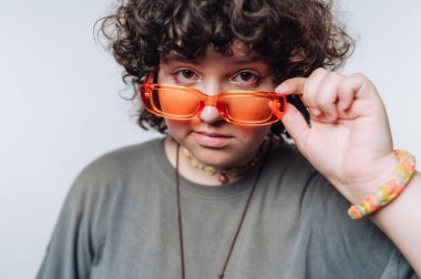 Açık arkaplanda parlak turuncu gözlük takan kıvırcık saçlı genç bir kadının portresi.