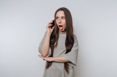 Genç bir kadının portresi bir telefon görüşmesinde şaşkınlıkla tepki verir, hafif arka planda duygu ve merakın bir karışımını gösterir.