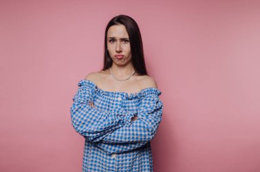 Suratı asık genç bir kadının portresi çapraz kollarla poz veriyor, pembe arka planda mavi kareli bir kıyafet giyiyor.