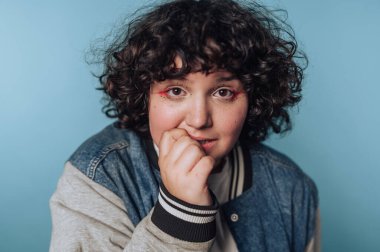 Düşünceli, kıvırcık saçlı genç bir kadının portresi parmaklarını ısırıyor, mavi arka planda duygularını gösteriyor.