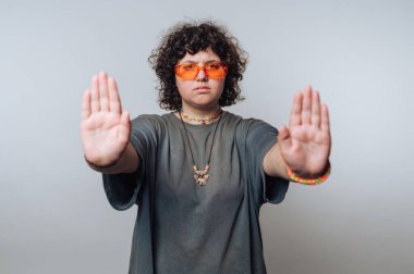 Şaşırmış, turuncu gözlüklü, kıvırcık saçlı genç kadının portresi açık arkaplanda kameraya el hareketi gösteriyor.