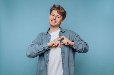 Genç bir adamın portresi, açık mavi arka planda dururken sevgi ve neşe taşıyan elleriyle bir kalp şekli oluşturur.