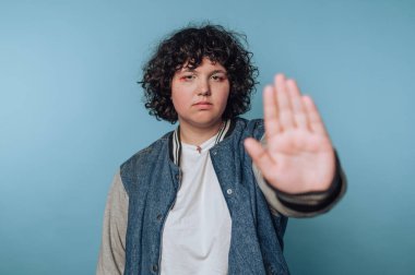 Genç bir kadının portresi, mavi arka planda ciddi bir ifade sergileyerek, dur işareti yaparak ellerini kaldırır.