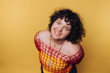 Kıvırcık saçlı genç bir kadının portresi canlı bir kıyafet sergiliyor ve sarı arka planda neşe yayıyor.