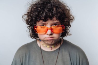 Turuncu güneş gözlüklü, açık arkaplanda ciddi bir ifadeyle şaşırmış kıvırcık saçlı bir kadının portresi.
