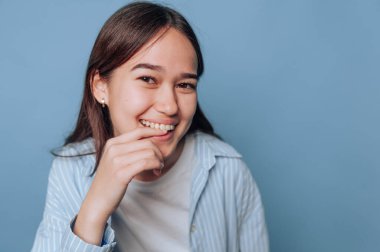 Genç kızın portresi utangaç bir şekilde gülümseyerek çenesine dokunuyor, açık mavi arkaplanda ışıldayan cazibe ve sıcaklığı.