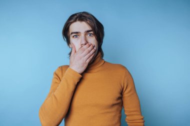 Genç adamın portresi eliyle ağzını kapatıyor, mavi arka planda şok ya da inançsızlık belirtileri gösteriyor.