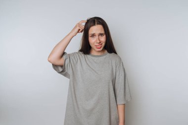 Gri tişört giymiş genç bir kadının portresi şaşkın bir şekilde duruyor, açık arka planda kafasını kaşıyordu.